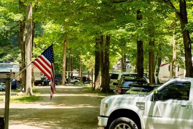Driveway/road through wooded RV sites showing vehicles and flags — illustrates the seasonal/long-term camping area and campsite layout. From the Seasonal Camping page.