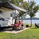 People relaxing beside a white RV under an awning by a lakeside campsite