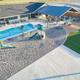 Aerial view of a pool area with a swimming pool, a playground with slides and climbing structures, a shaded pavilion with seating, and an adjacent grassy area.
