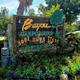 Bayou Campground -A rustic wooden sign reads "Bayou Campground, 5001 Hwy 3, 77539, Dickinson, TX," surrounded by lush plants and greenery.