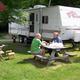 Campsite scene with a travel trailer, picnic table and guests seated — clear photo of an individual RV pitch and typical guest area; appears on the campground homepage/rates pages.