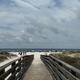 A wooden boardwalk leads over sand dunes to a wide beach lined with colorful umbrellas and beachgoers. The sky is mostly cloudy, and the ocean is visible in the distance.