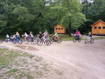 Shot of a bike parade with small cabins visible in the background and campsite road — demonstrates campground layout, activities and cabin accommodations.