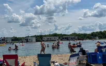 Beach/lake scene with people and RVs in the background — clearly shows the on-site swim/rec area and shoreline activity referenced by the site images.