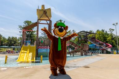 Wide view of the pool and water-play complex with slides and mascot in the foreground — strong depiction of the park’s aquatic facilities and family amenities.