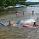 Shallow beach area with children swimming, flotation noodles and dock/boats — depicts the campground’s swimming/beach area and family activities (source: Facilities page).