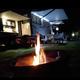 Nighttime camping scene with a lit campfire in the foreground, a recreational vehicle (RV) illuminated with interior and exterior lights, equipped with a canopy, and a picnic table with a tablecloth and chairs.