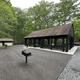Opened sided, roofed picnic pavilion in the forest near a flat surface with two standing grills with a restroom building nearby