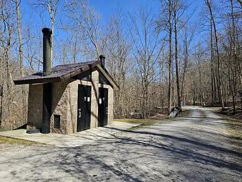 Vault toilet facility sits in gravel parking lot of campground.