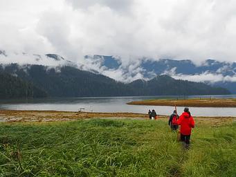 Harding River Cabin scenery, four people walking along grassflats to a boat with a river and clod covered mountains in background