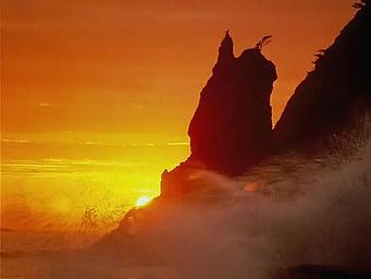 A dramatic closeup picture of a sea stack at sunset. Waves crash on the sea stack and the sunset colors a bright orange gradient in the sky.