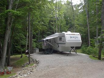 Gravel RV site with a fifth-wheel camper and awning in a wooded setting — good close-up of typical RV pitch, from Amenities page.