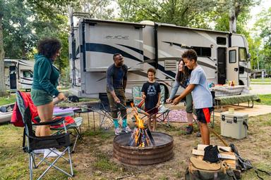 Clear photo of an RV site with a family around a fire ring and an RV in the background — demonstrates typical RV pitch layout and amenities (fire ring, chairs), matching the site’s ‘RV sites’ gallery context.