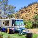 An RV is parked on a green mat with chairs and a fire pit. It’s surrounded by hills and trees under a clear blue sky.