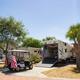 A golf cart with an American flag is parked near a large RV with its back ramp open in a sunny, paved camping area with trees and palm trees.