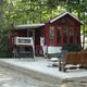 Cabin with large front porch overlooking a pond, surrounded by trees, outdoor patio with metal table and chairs, a wooden bench, and a small fire pit.