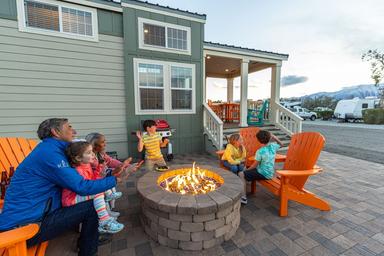 Image of a family gathered around a firepit on a paved patio by a cabin structure with RVs visible in the background — illustrates cabin lodging and shared outdoor amenities at the campground (from the site’s waystostay photos).