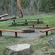 Large flat rock in front of benches around a fire ring with picnic table, grassy meadow, and forest in background.