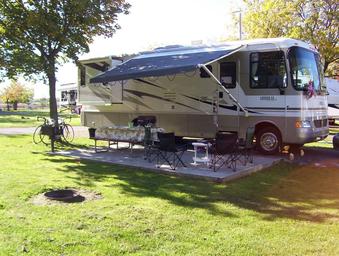 Close view of an RV on a concrete patio with awning, picnic table and grassy site area — clearly depicts full-hookup RV campsite amenities available at the KOA.
