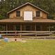 House with shingle siding,  solar panel on the roof of wrap around covered porch, hand pump well on the lawn, and ramp leading up to porch