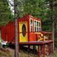 A small, colorful house on stilts in a wooded area, with a red exterior, yellow door, and yellow porch furniture. A person is relaxing in a hammock beside the house.