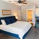Bedroom with a large bed, blue accents, two chairs, a ceiling fan, lamps, a wall-mounted TV, and a glimpse of a bathroom in the background.