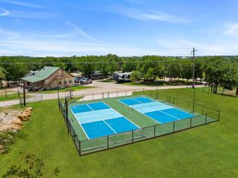 Aerial view of two blue tennis courts beside a grassy field and small building under a clear sky