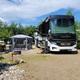 Wide view of a large RV parked at a leveled gravel/KOA-style site with patio, fire ring and picnic setup — concrete evidence of RV accommodations on the property.