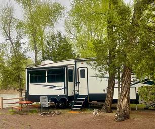 River-site showing a large trailer parked near trees and the riverbank — another clear example of typical riverfront RV/campsite on the property (from homepage/gallery).