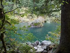 From a rocky shore in the shade of the forest looking at a moss flecked boulder in a deep jade river.