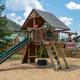 A wooden playground with slides, climbing ladders, a tire swing, and a shaded fort, set among trees under a blue, partly cloudy sky.