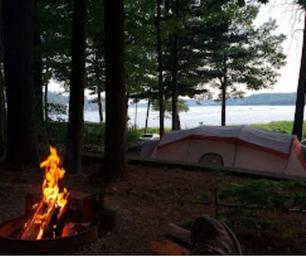 Tent campsite at the water’s edge with a campfire ring and lake visible through trees — illustrates tent camping and waterfront sites (used on Camping Fees page).