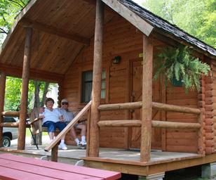 Close view of a rustic cabin with a covered porch and guests seated — useful for showing available cabin lodging and porch amenities on the property’s listing.