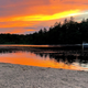 Sunset over a calm lake reflecting orange sky, with dark trees along the shore and a sandy beach in front