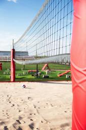 Beach volleyball court with a volleyball on the sand, visible net, and a red padded post support.