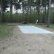 Concrete RV/trailer pad with picnic table and surrounding pine trees — shows an individual RV/campsite layout available at Sandy Pines (Camping page / site photos).