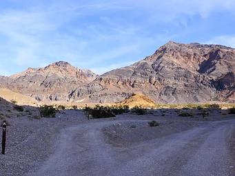Gravel dirt road with parallel roadside campsite shaded in the foreground, sunny mountains in the background.