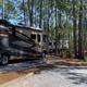 Close view of a motorcoach parked at a gravel site among pine trees — shows RV pitch layout, hookups and site setting.