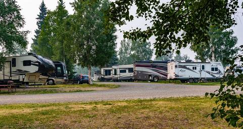 Wide banner view showing a row of parked RVs at the site with grassy foreground and trees — useful campsite/RV pitch scene demonstrating layout and unit size.