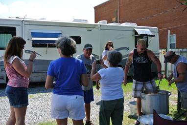 Photo of people gathered next to a parked RV on a gravel site with a brick building in background; appears to show on-site RV campsite/activity and matches the site’s social image (blob-0008.png) on the property pages.
