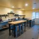 Communal kitchen with stainless steel counter, dish shelves, microwave, and butcher-block high tables with blue metal stools.