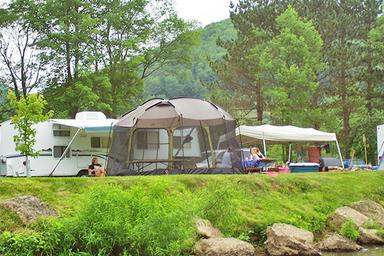 Riverside campsite with tent, trailer and lawn area adjacent to rocks/riverbank — clearly shows tent/RV camping options (from the site’s Rates & Reservation gallery).