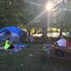 Tent campsite with canopy of trees, picnic table and people relaxing — illustrates tent camping area and pond/wooded setting (from 'ways to stay' imagery).
