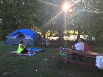 Tent campsite with canopy of trees, picnic table and people relaxing — illustrates tent camping area and pond/wooded setting (from 'ways to stay' imagery).
