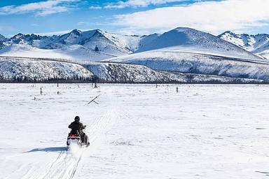 A snowmobile riders crosses a big meadow with mountains in the distance.