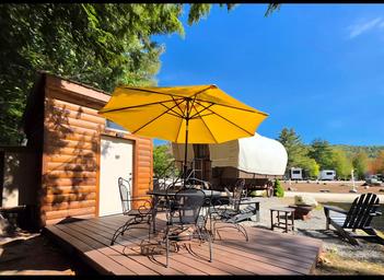 Deck and raised cabin structure set among trees with Adirondack chairs and nearby RVs visible — useful photo of the campground’s cabin lodging and wooded setting.
