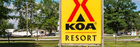 Virtual tour thumbnail from the Port Huron KOA page showing an on‑site scene with RVs, guests, and cornhole games — depicts the campground layout and recreational area.