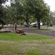 Small photo of a grassed campsite area with picnic table, fire ring and adjacent paved pad — useful close-up of a typical campsite. Found on the About / Camping Rates context (about.html), supporting relevance to the property.