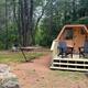 Photograph of a small wooden hex-style cabin on a cleared campsite with picnic table, hammock and surrounding trees — directly shows cabin lodging offered on the property (file hosted on the campground site).