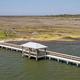 Aerial view of a long wooden fishing pier with two covered gazebos extending from a marsh shoreline into calm water.